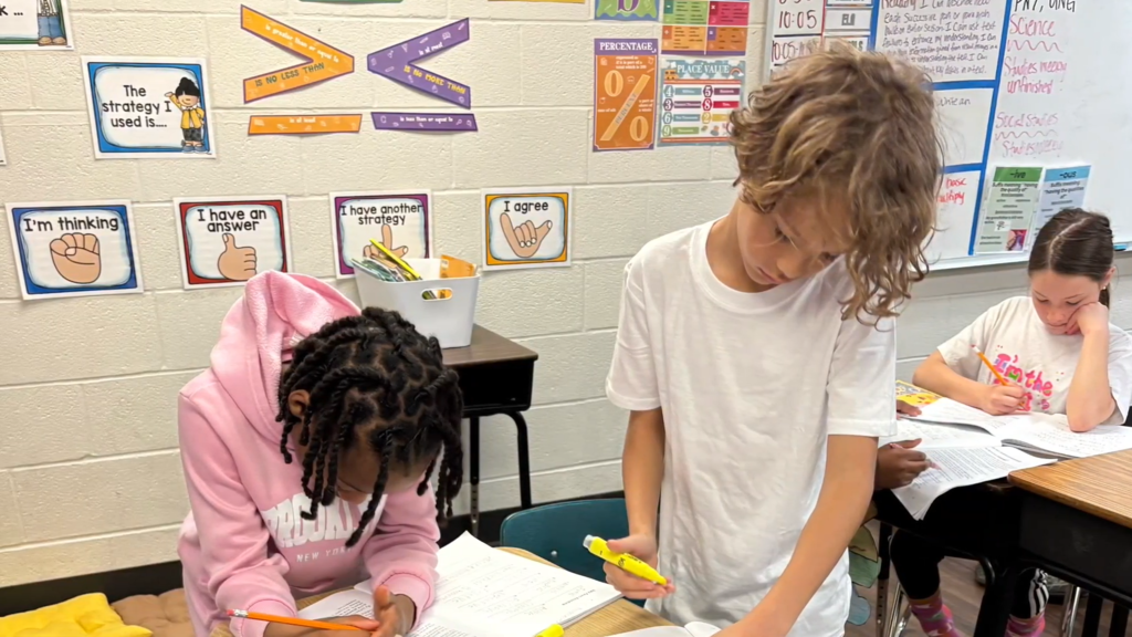 Three students working on assignments in a classroom. A boy stands holding a yellow highlighter while two girls sit at their desks writing in their workbooks.
