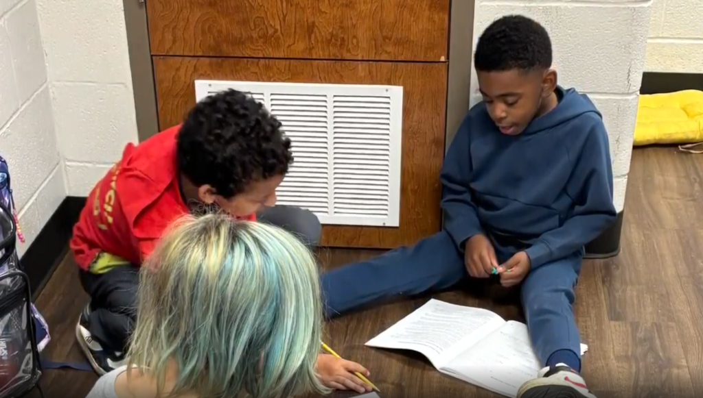 Three students sit on the floor. One is in a red shirt and another in a blue hoodie. They are working together on an open workbook.