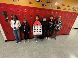 Students holding up a building they built out of cardboard. They are standing shoulder to shoulder with lockers behind them.