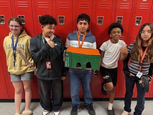 Students holding up a building they built out of cardboard. They are standing shoulder to shoulder with lockers behind them.