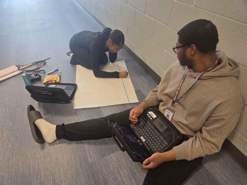 Student writing on a piece of paper on the ground as she researches from a laptop. Another students is researching beside her.