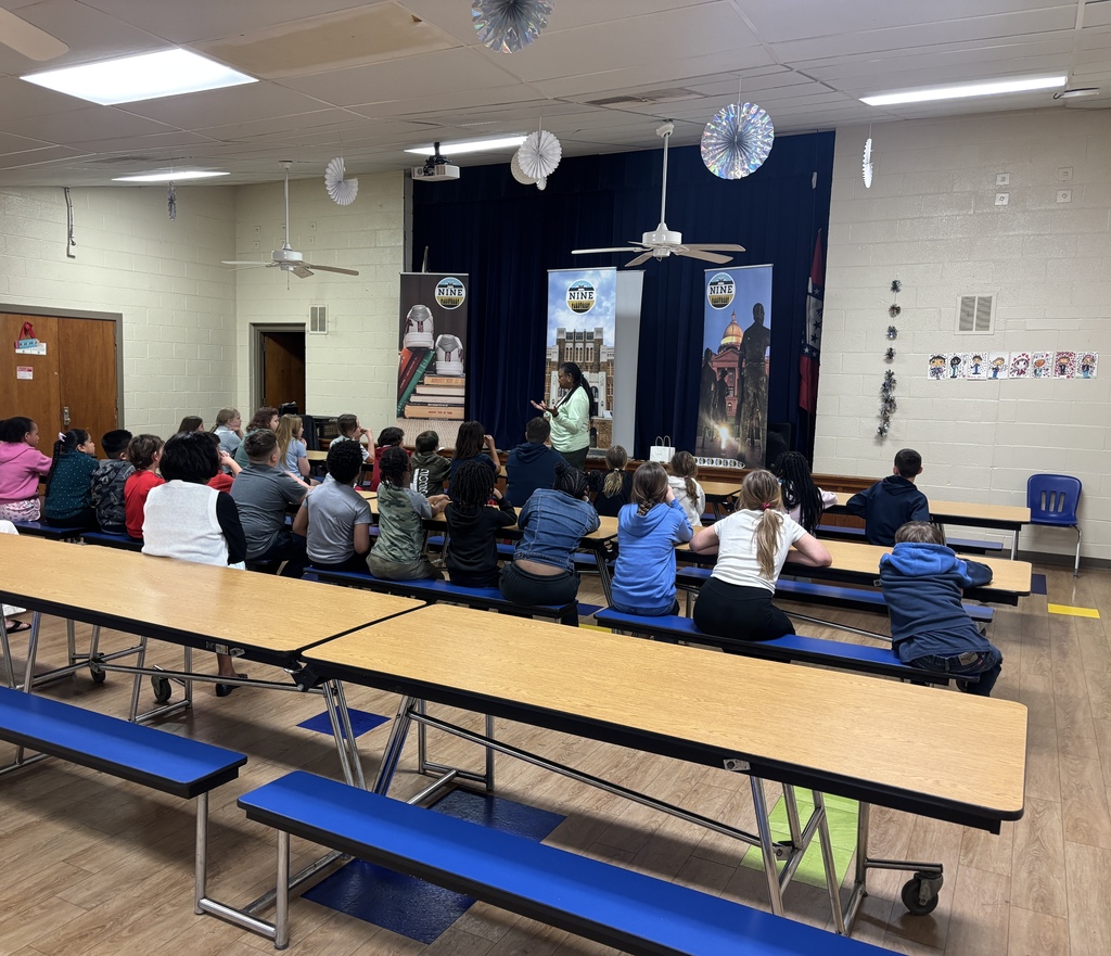 Image of Agnolia B. Gay in front of 4th grade students and teachers. She is educating the group on the Little Rock Nine. There are three banners behind her. One with books on it., one with Little Rock Central High National Historic Site, and one with the Little Rock Capitol Building. 
