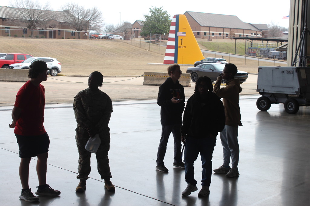 Six people stand silhouetted inside a hangar opening, looking out toward an aircraft tail marked with numbers and an American flag, with vehicles and buildings visible outside.