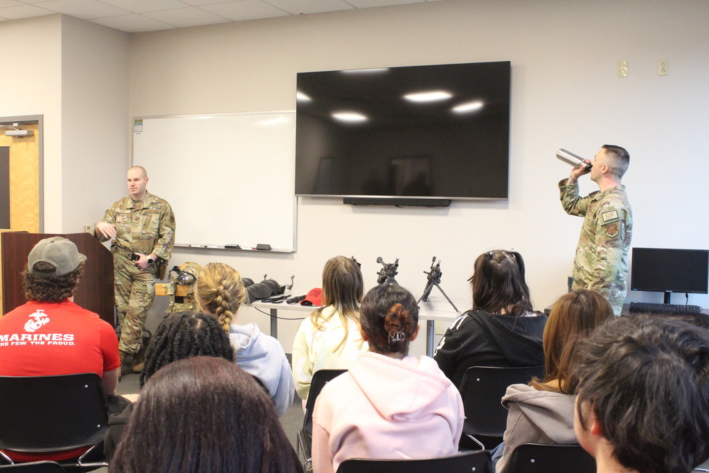 Two military presenters stand at the front of a classroom, one speaking at a podium while the other drinks from a metal container; military gear and equipment are displayed on the table in front of them as students listen.