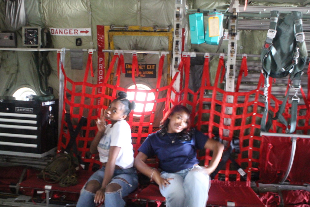 Two people sit on red webbed seats inside a military aircraft, posing for a photo with safety equipment, storage containers, and an emergency exit door visible behind them.