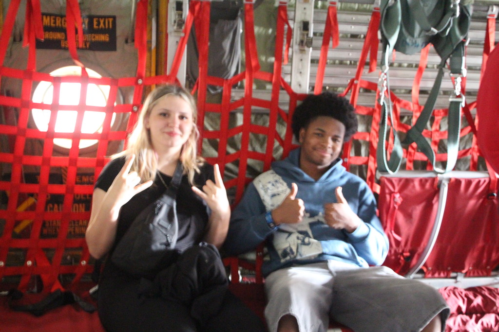 Two individuals sit inside a military aircraft on red webbed seating, smiling and gesturing toward the camera, with an emergency exit window and aircraft interior components behind them.