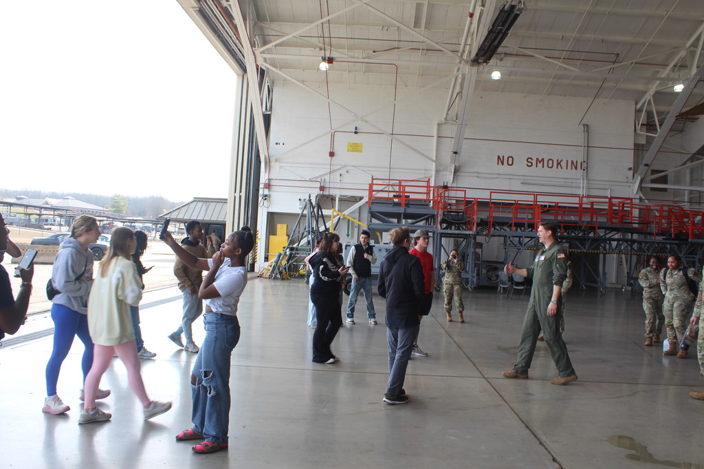 A group of civilians and military personnel stand inside a large aircraft hangar, talking and taking photos near industrial equipment and a red metal platform structure.