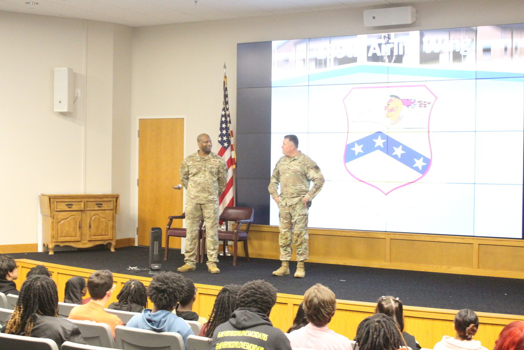 Two military personnel in uniform speak to a group of students seated in an auditorium-style classroom, with a large screen behind them displaying a military insignia.