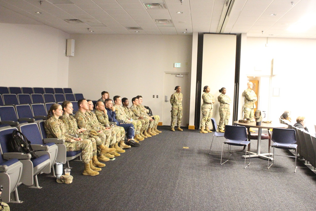 A room full of military personnel in camouflage uniforms sit in rows of blue chairs during a briefing or training session, with a few individuals standing along the back wall.