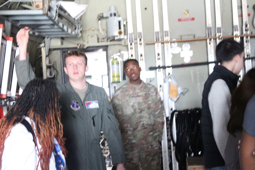 A group of people, including a pilot in a green flight suit and a service member in camouflage, stand inside a military aircraft while civilians observe the equipment and interior structure.
