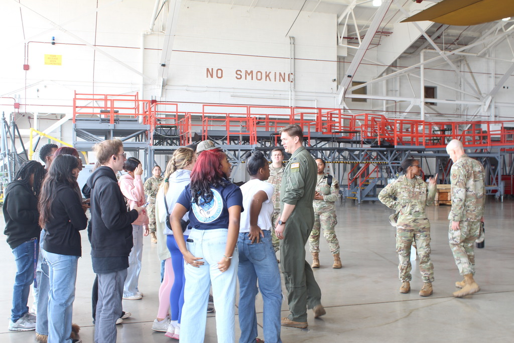 Civilians and military personnel gather inside an aircraft hangar, conversing near industrial platforms and staircases, with a large “NO SMOKING” sign visible on the wall.