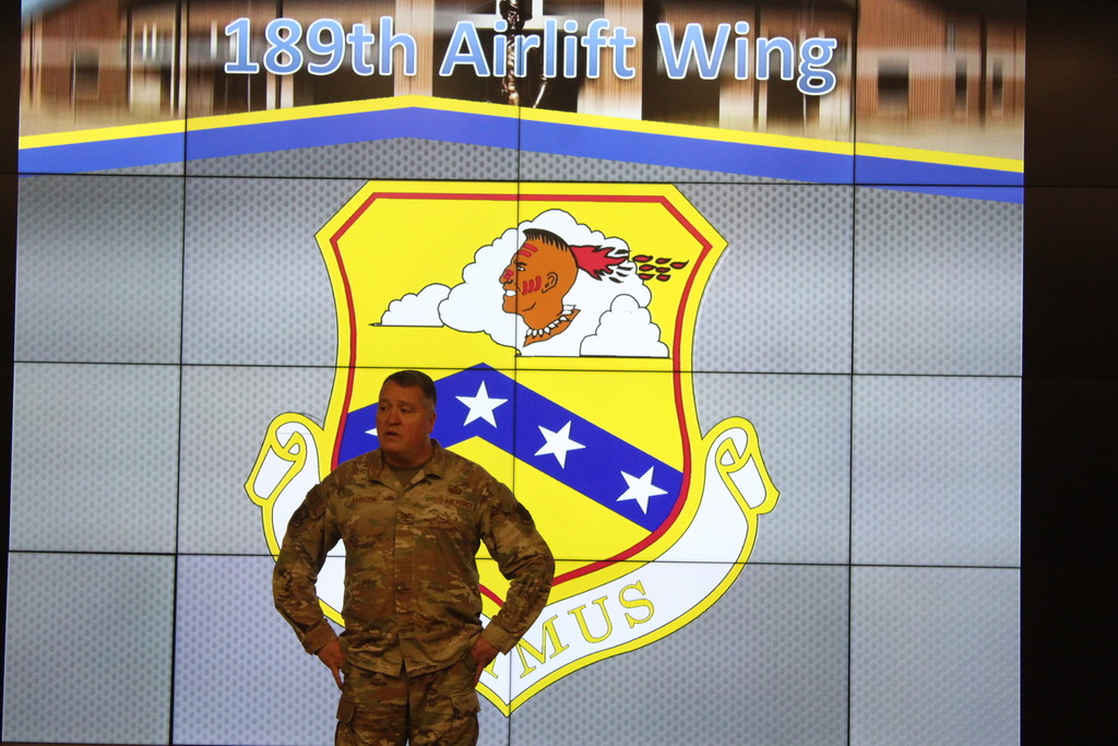 A U.S. Air Force service member stands in front of a large digital display showing the emblem and name of the 189th Airlift Wing.