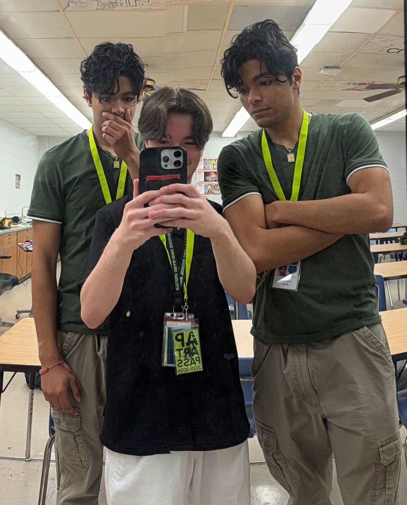 Three students take a mirror selfie in a classroom; the student in the center holds the phone while the two on either side, dressed alike, appear as twins, with classroom desks and decorated ceiling tiles in the background.