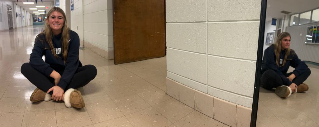 A student sits cross‑legged in a hallway near a corner mirror, which reflects their image in a way that has an opposite expression.