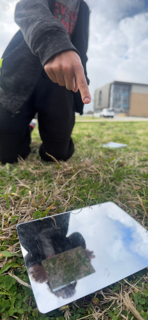 A student kneels on grass outdoors, pointing at a small mirror on the ground that reflects the sky and part of their hand, with a modern building visible in the background.