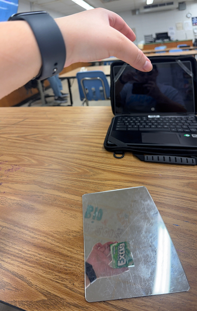 A mirror placed on a classroom desk reflects a hand holding a pack of gum and reversed wall text, while a laptop and blue classroom chairs sit around it, creating a reflection‑based illusion.