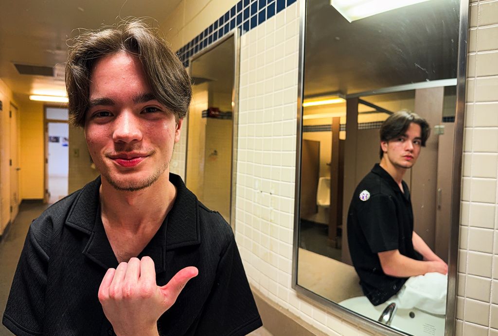 A student stands in a restroom pointing toward a mirror that reflects them sitting on a sink, creating a visual illusion of two different positions of the same person within the same space.