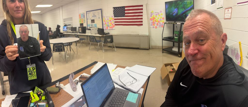 In a classroom, a student holds a small mirror that reflects the face of another person seated at a desk with a laptop and papers, creating a playful optical illusion using the mirror’s angle and a different expression.