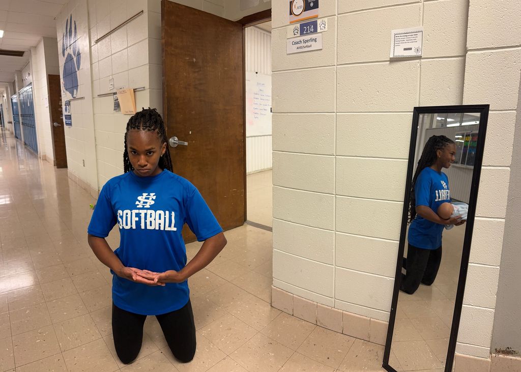 A student kneels in a school hallway with a serious expression, while a mirror beside them reflects a smiling version of the same student holding a baby doll, creating a contrast between the real pose and the reflection.