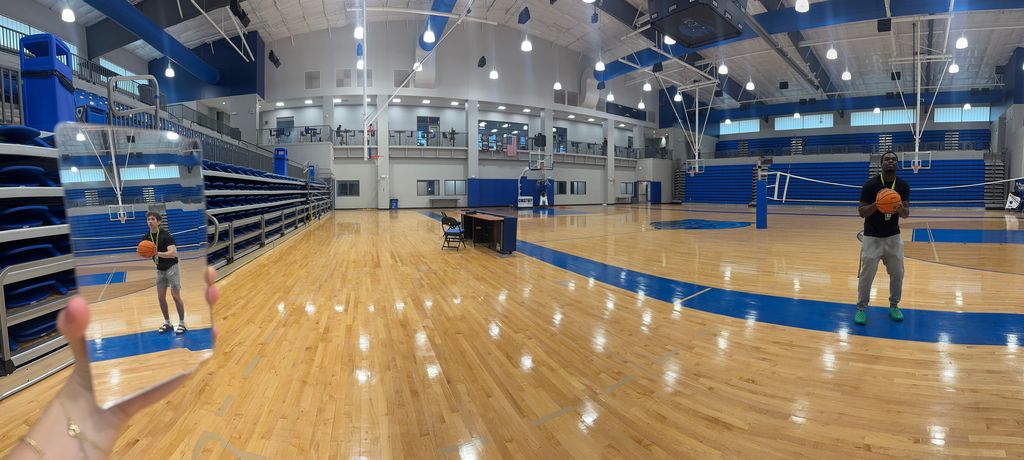 Inside a gymnasium, a person holds a mirror that reflects another individual holding a basketball, creating the illusion that the reflected person is present in the gym even though they are not physically in the frame.