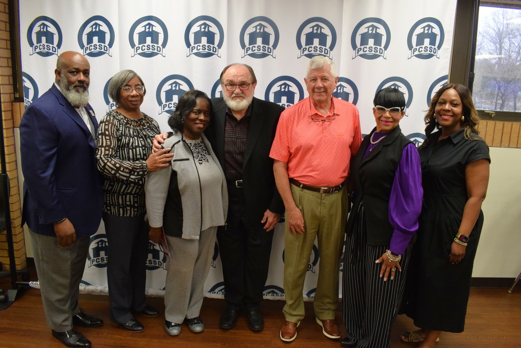 Seven adults pose together in front of a step‑and‑repeat backdrop featuring the PCSSD logo. They are dressed in business attire and smiling for a group photo.