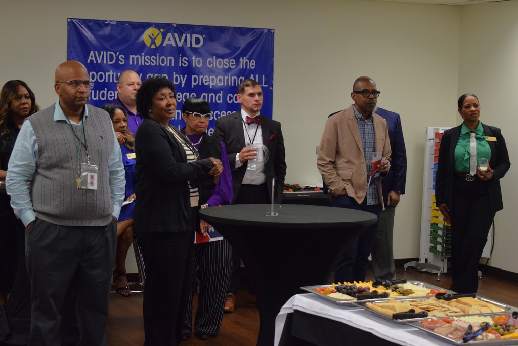 A group of professionally dressed adults stand around a tall table and a buffet spread with fruit, cheese, and crackers during a reception. A large blue AVID banner with the program’s mission statement is displayed in the background.