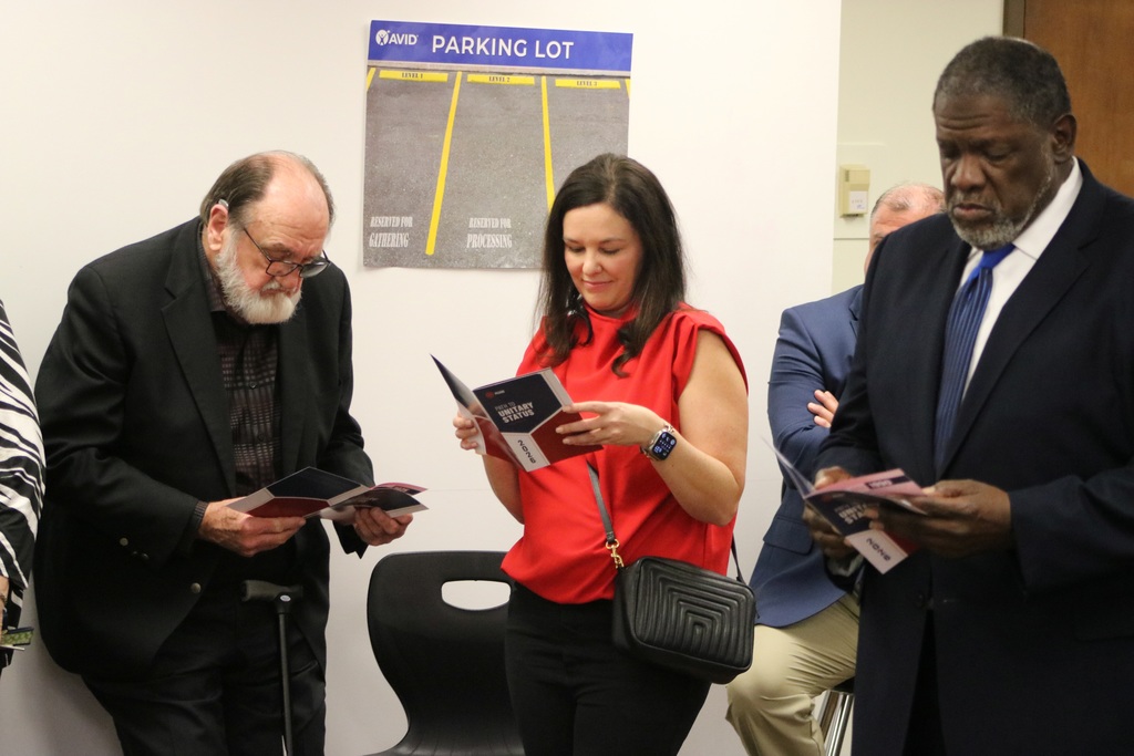 Several adults stand indoors reading pamphlets during a professional development session. A poster on the wall labeled “AVID Parking Lot” displays three levels with reserved spaces for gathering and processing.
