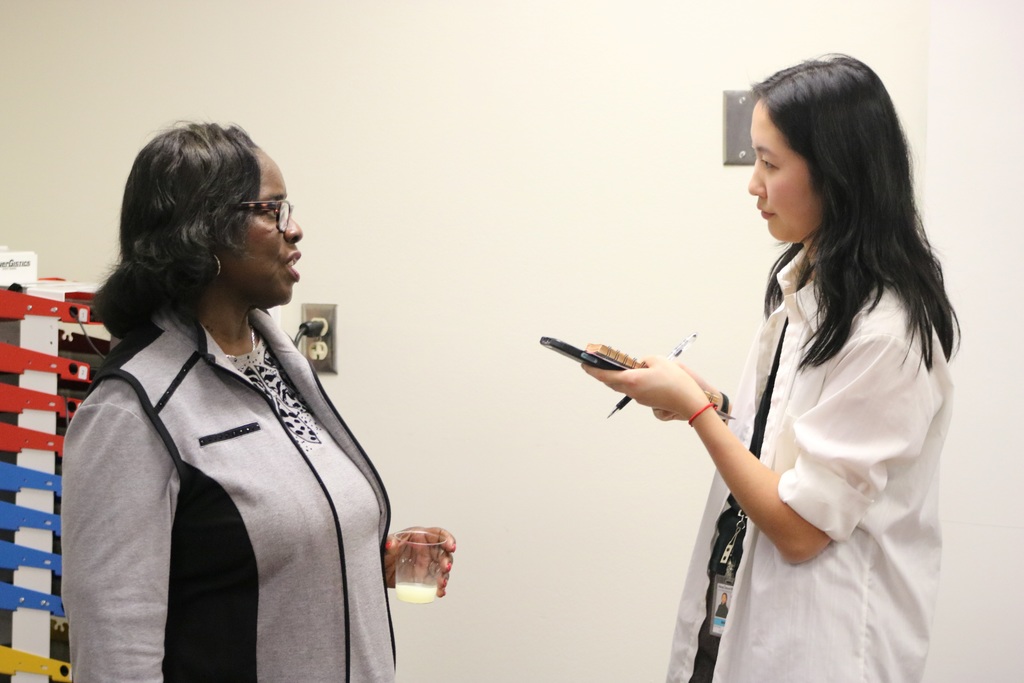 A reporter holding a notepad, phone, and pen interviewing another woman holding a glass.