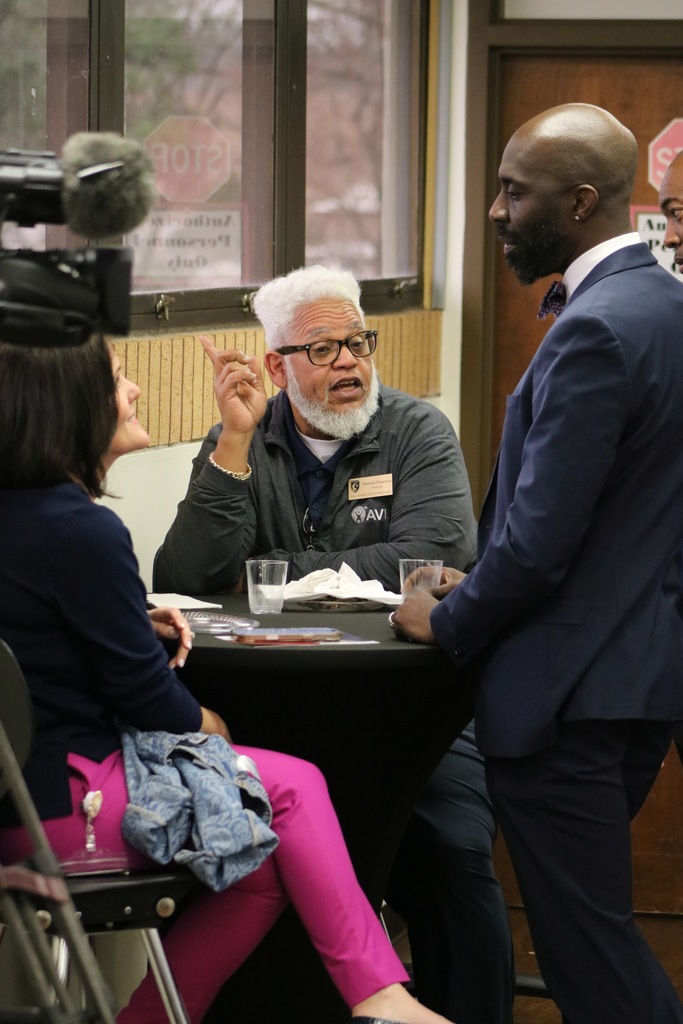 Three adults engage in a conversation around a small table while a camera with a microphone records above them. One person gestures with a raised finger, and another stands listening attentively.