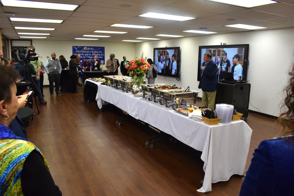 Guests mingle around a long buffet table filled with catered food at a formal reception. A speaker stands at a podium with two display screens behind him showing a group photo. A blue AVID mission banner hangs on the wall.
