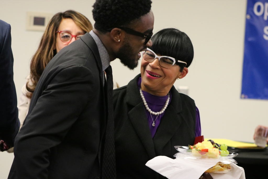 A man in a suit leans toward a woman holding a plate of fruit as they talk during a reception. Another woman stands behind them. A blue banner with partially visible text hangs on the wall.