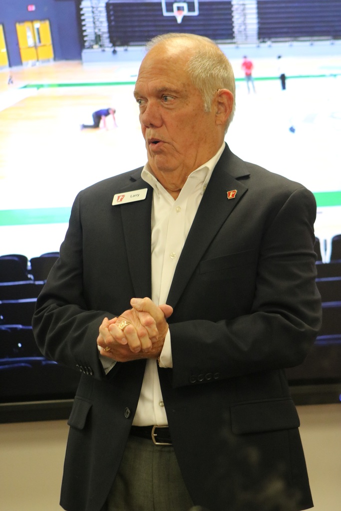 A man wearing a suit jacket and a name tag reading “Larry” stands indoors with his hands clasped, appearing to speak. Behind him, a large screen shows an image of a gymnasium with people in the stands.