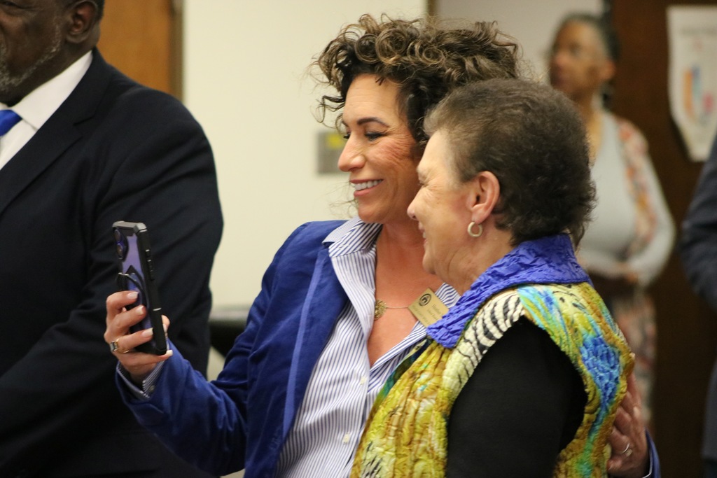 Two people stand close together smiling at a smartphone held by the person on the left. They appear to be taking or viewing a photo during a social gathering, with other attendees visible in the background.