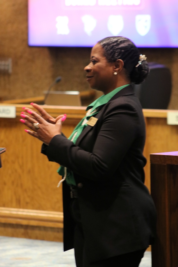 A PCSSD board member stands in the boardroom wearing a formal black outfit with a green shirt and name badge, appearing to clap or gesture during the recognition.