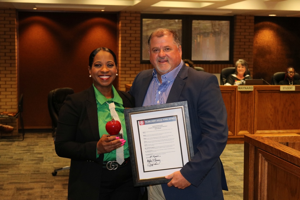 An outgoing PCSSD board member stands with Superintendent Jeff Senn holding their awards—one with a red apple trophy and the other with a framed resolution—during the board meeting recognition.