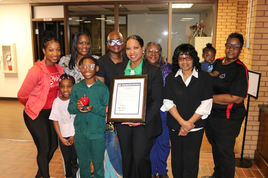 A group of ten people, including adults and a child, pose together after the recognition of an outgoing PCSSD board member. One person holds a framed resolution, and the child holds a red apple‑shaped award.
