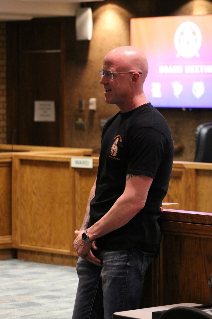 A PCSSD board member stands at the front of the boardroom during the meeting, wearing a black T‑shirt and glasses. A digital “Board Meeting” screen is visible behind him.