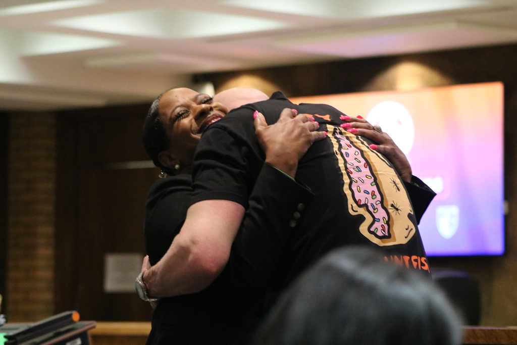 Two PCSSD board members embracer in a warm hug during the recognition resolution. A digital meeting screen and boardroom seating are visible in the background.