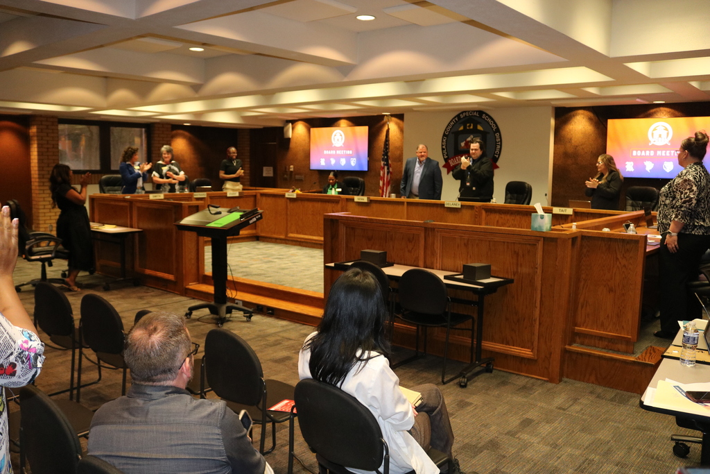 Board members and attendees stand and applaud inside the PCSSD boardroom during a recognition moment. The room features a U‑shaped board table, digital “Board Meeting” screens, and the district seal on the wall.