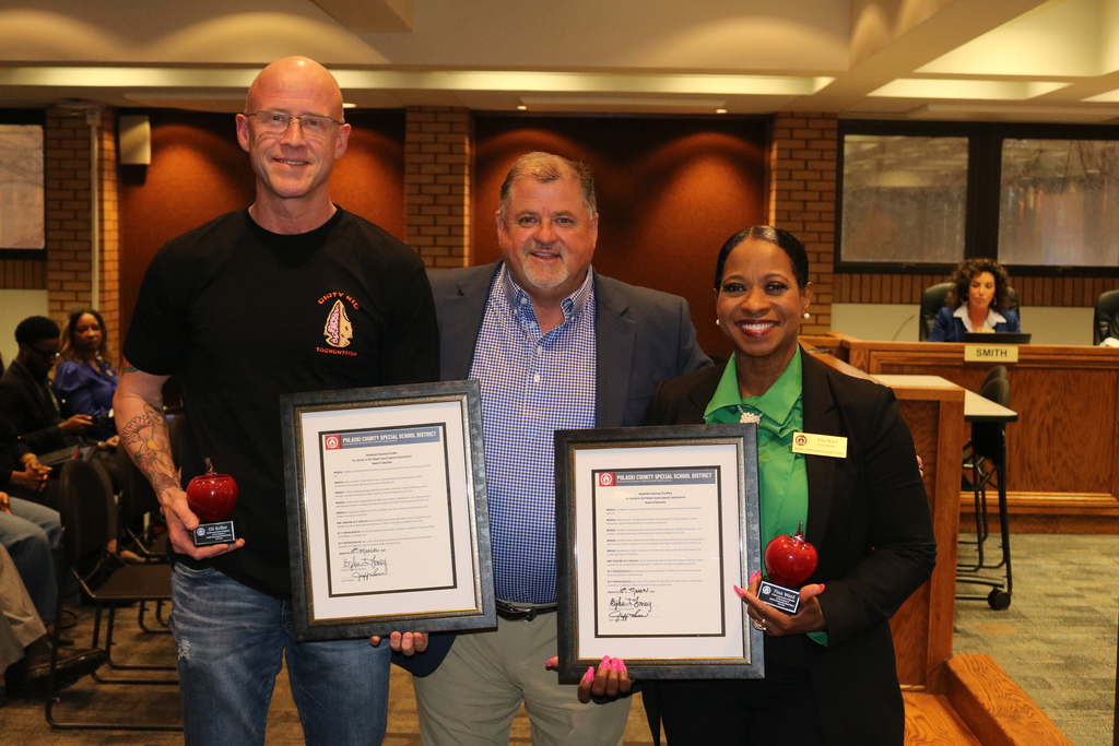 Two outgoing PCSSD board members stand on either side of Superintendent Jeff Senn, each holding a framed Resolution for Service and a red apple‑shaped award. Other attendees are seated behind them.