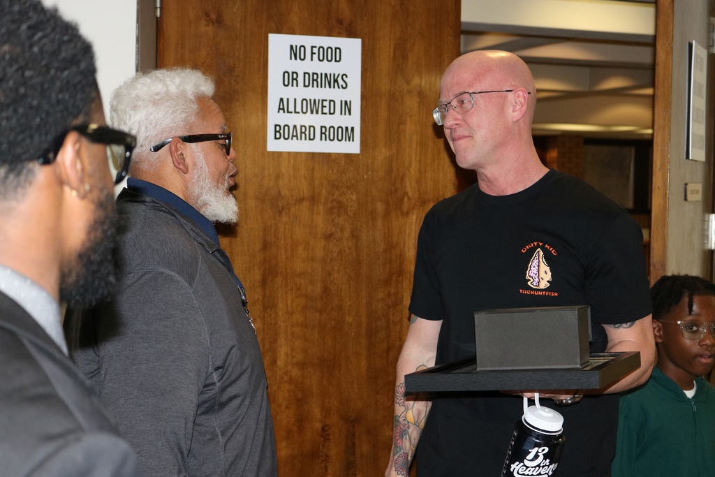 Two individuals converse near the entrance to the PCSSD boardroom. One holds a picture frame with items while standing beside a sign that reads “No Food or Drinks Allowed in Board Room.”