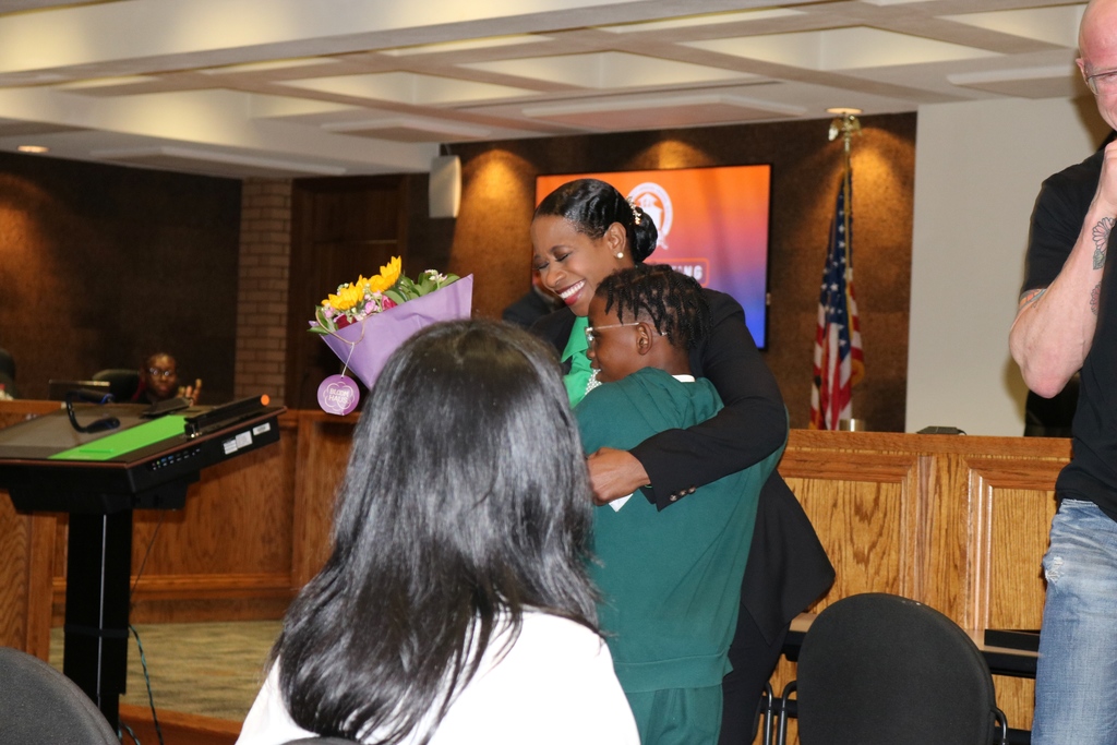 A PCSSD board member hugs a child holding a bouquet of purple‑wrapped flowers during the recognition ceremony. A podium, digital meeting screen, and American flag are visible in the background.