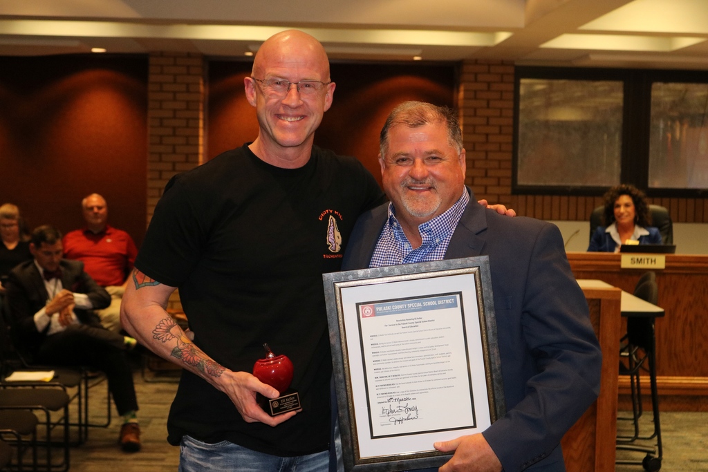 An outgoing PCSSD board member stands with Superintendent Jeff Senn holding their awards—one with a red apple trophy and the other with a framed resolution—during the board meeting recognition.