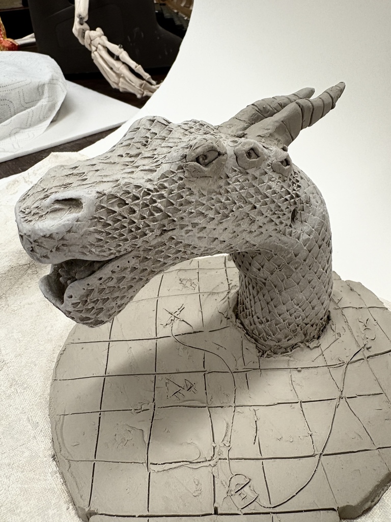 A detailed clay sculpture of a dragon’s head and neck mounted on a circular base. The dragon has textured scales, curved horns, and an open mouth. The base is carved with grid-like patterns and symbols. Art supplies and a skeletal arm model sit in the background.