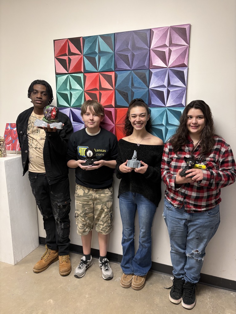clay busts 7our students stand in front of a colorful geometric mural made of raised diamond-shaped panels. Each student holds a small art piece, and they stand on a concrete floor beside a white wall.