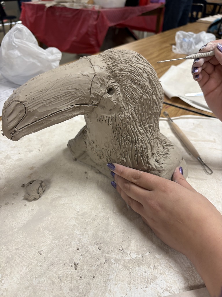 A student uses a metal sculpting tool to carve details into a clay toucan sculpture. The bird’s beak and feathers are partially shaped, and several sculpting tools lie on the worktable.