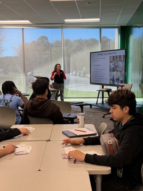 A presenter stands at the front of a classroom speaking to students seated at tables. A large screen behind her displays a slide titled “Fighting Over Social Fit.”