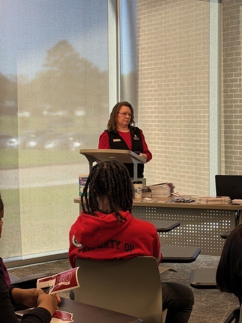 Students sit in rows of modern classroom chairs with attached desks, listening to a presentation. Large windows line one side of the room, letting in natural light.