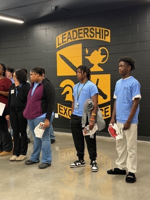 A group of students stand in front of a black wall featuring a large yellow graphic with the words “Leadership” and “Excellence.” Several students hold booklets.