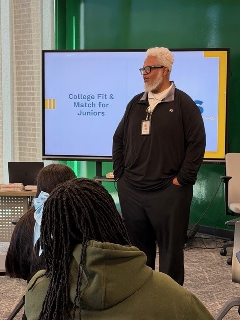 A presenter stands at the front of a classroom speaking to students seated at tables. A large screen behind her displays a slide titled “College Fit & Match for Juniors".”